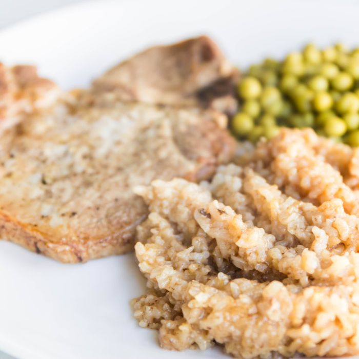 pork chop, seasoned rice, and peas on a white plate on table 